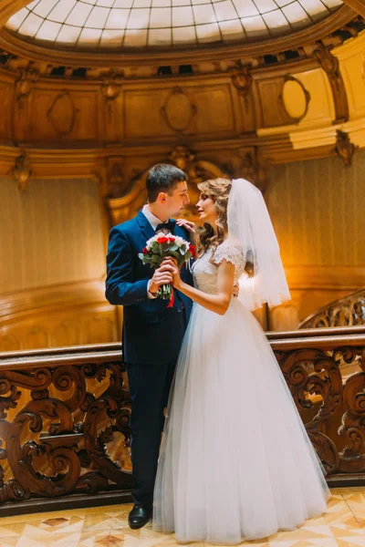 Encantadora novia y novio elegante guapo cerca de balaustres de madera vintage con el fondo del interior de lujo —  Fotos de Stock