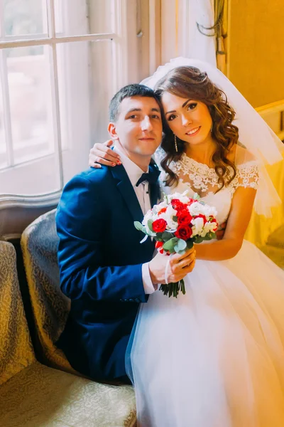 Enloved ζευγάρι ποζάρει κοντά σε παράθυρο φόντο με το εκλεκτής ποιότητας εσωτερικό. Νύφη στο γάμο φόρεμα κάθεται στα γόνατά της τον γαμπρό της — Φωτογραφία Αρχείου