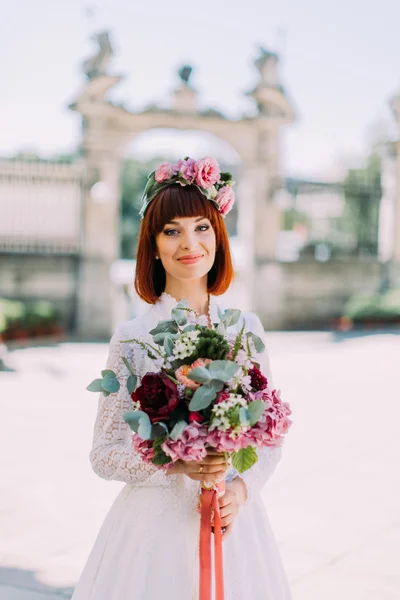 Hermosa novia pelirroja con corona de flores al aire libre de la ciudad en el día soleado. Boquet en mano . — Foto de Stock