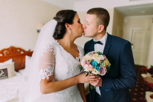 Splendida sposa ed elegante sposo baciare in camera d'albergo dopo la cerimonia di nozze. Sposi novelli in luna di miele — Foto Stock