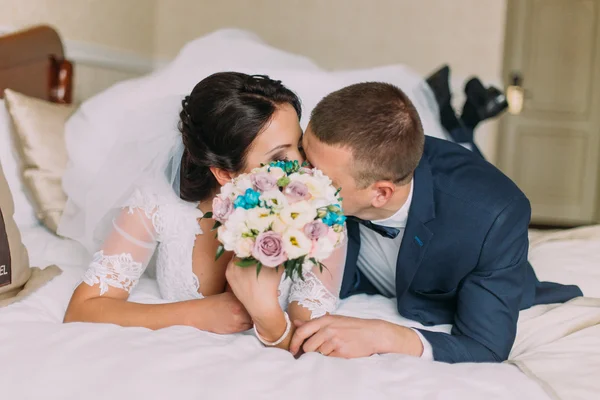 Feliz recién casados yacen en la cama en la habitación de hotel después de la celebración de la boda y compartir beso —  Fotos de Stock