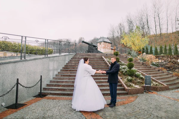 Красива наречена в довгій білій сукні позує з елегантним нареченим біля сходів на відкритому повітрі. Стара гармата на фоні — стокове фото