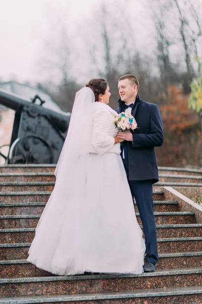 Красивая невеста в длинном белом платье позирует с элегантным женихом на лестнице на открытом воздухе. Старая пушка на заднем плане — стоковое фото