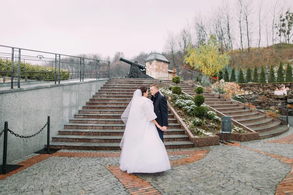 Красива наречена в довгій білій сукні позує з гарним нареченим біля сходів на відкритому повітрі. Стара гармата на фоні — стокове фото