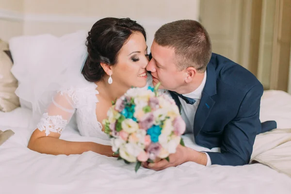 Feliz cansado recién casados yacen en la cama en la habitación de hotel después de la celebración de la boda y compartir beso —  Fotos de Stock