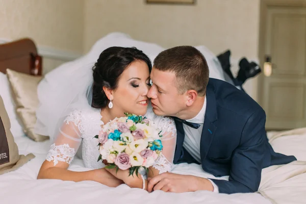 Ευτυχισμένη νύφη και τον γαμπρό βρισκόταν στο κρεβάτι στο δωμάτιο ξενοδοχείου μετά γαμήλια γιορτή. Νεόνυμφοι τρυφερά φιλιά — Φωτογραφία Αρχείου