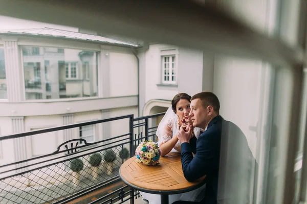 La sposa splendida e lo sposo elegante si siedono al tavolo su balcone di camera d'albergo. Sposi luna di miele concetto — Foto Stock