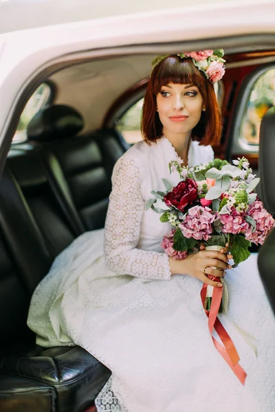 Olhe através das portas do carro aberto em uma noiva de cabeça vermelha deslumbrante com buquê rosa de flores — Fotografia de Stock