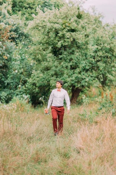 Noivo feliz com buquê de casamento caminhando para sua noiva através do prado da floresta de verão — Fotografia de Stock