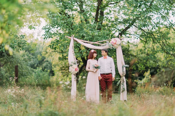 Casal vestindo trajes de casamento com buquê de flores e vegetação no jardim, arco no fundo — Fotografia de Stock