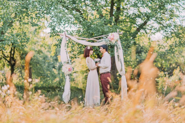 Стильна щаслива наречена і наречена обіймаються, дивлячись один на одного на весільну церемонію в саду біля арки з квітами — стокове фото