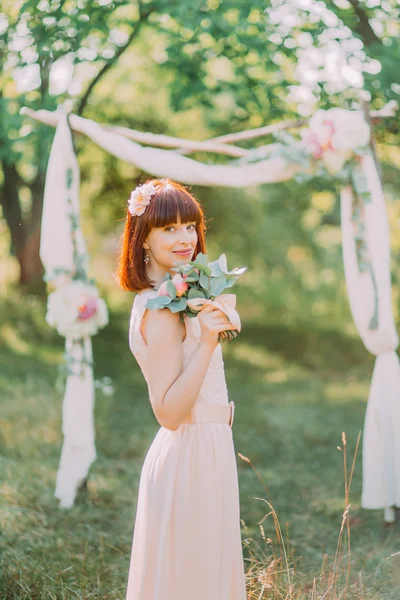 Вид збоку огидна руда щаслива наречена в білій сукні позує в парку, прикрашеному квітами — стокове фото