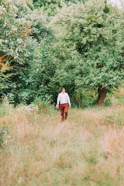 Jovem de terno elegante andando na trilha por campo de verão com buquê. Floresta no fundo — Fotografia de Stock