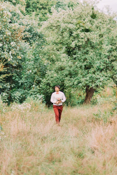 Elegante elegante jovem noivo em calças cor de tijolo com buquê de flores andando sozinho no parque — Fotografia de Stock