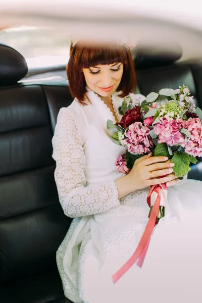 Preciosa novia con hermosa sonrisa está sentado en el coche de la boda y la celebración de flores de verano ramo — Foto de Stock