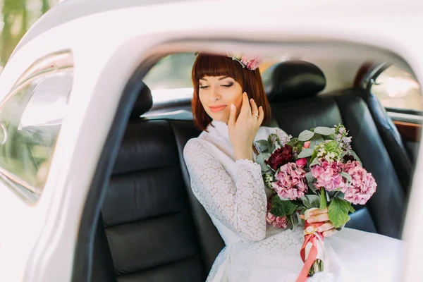 Día de la boda. Novia sentada en el coche, mirando hacia abajo, sonriendo y sosteniendo el ramo — Foto de Stock