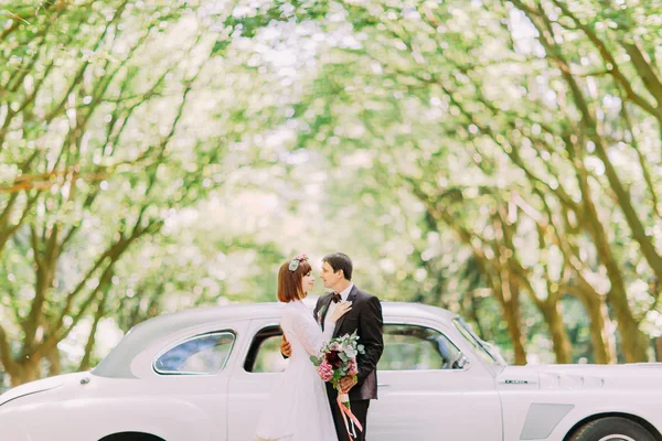 Bela noiva ruiva e elegante noivo abraçando perto de carro retro elegante sob a luz do sol — Fotografia de Stock
