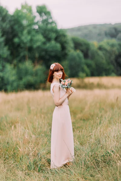 Full-length van sensuele roodharige bruid in een roze jurk reputatie op weide boeket — Stockfoto