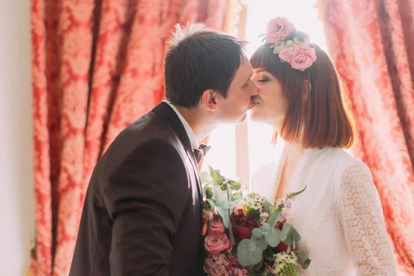 Romantisches Brautpaar küsst sich im Hotel in der Nähe des Fensters mit Blumenstrauß, Nahaufnahme — Stockfoto