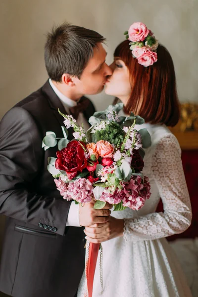 Charmante Braut im schönen weißen Kleid hält Strauß mit ihrem stilvoll gekleideten Bräutigam, sanft küsst — Stockfoto