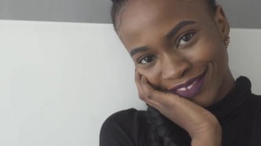 Gri arka planda çenenin altında el ile kameraya bakan zarif Afrikalı ya da siyah amerikalı kadının yakın çekimi