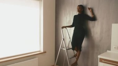 Koyu elbiseli güzel ve seksi afro-amerikan kadın stüdyoda gri duvara el yaslanmış uzakta görünümlü poz