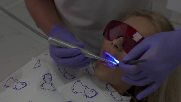 Paciente en cabitet dentista en tratamiento. Asistente médico oral utiliza luz azul primaria para endurecer compuesto dental — Vídeo de stock