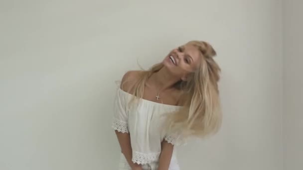 Attractive young blonde girl in white top laughs and plays with her hair in front of white plain wall — Stock Video