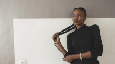 Charming ciddi afrikalı-amerikalı kadın uzağa bakıyor ve stüdyoda gri duvara yaslanmış saçlarına dokunuyor