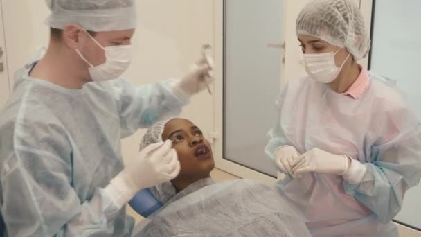 Vista laterale del dentista con assistente che indossa uniformi rendendo le procedure mediche paziente femminile in tazza scrup presso la clinica dentale — Video Stock