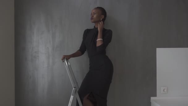 In voller Länge sexy afrikanische Frau in dunklem Kleid posiert wegschauend und hält die Hand unter dem Kinn an der grauen Wand im Studio — Stockvideo