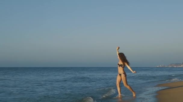 Bela jovem correndo para a água do mar na praia, salpicando e desfrutando de ondas costeiras. Movimento lento — Vídeo de Stock