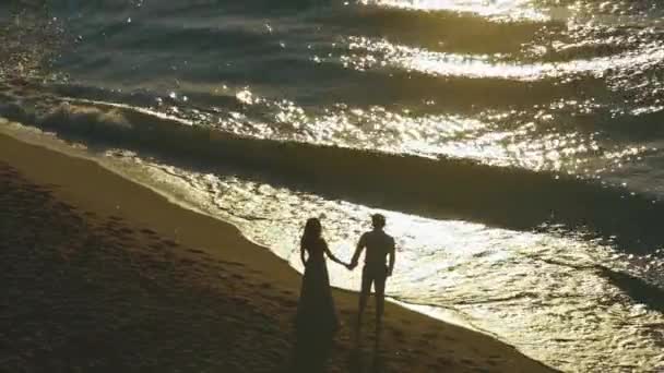 Silhuetas de par elvoved na praia da noite do por do sol. Casal olha como ondas quebrando na costa arenosa — Vídeo de Stock