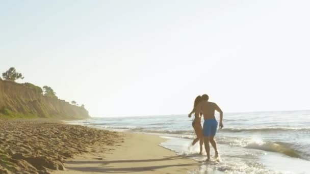 幸せな若いカップルは、砂浜のビーチでサーフィン波の海岸近く再生します。 — ストック動画