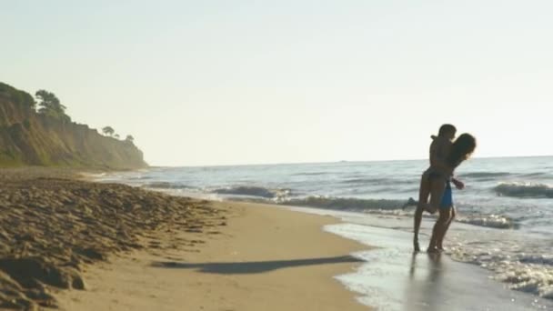 Joyeux jeune couple jouent près du bord de mer dans les vagues de surf sur la plage de sable fin. Beau homme tient jolie fille — Video