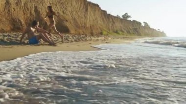 Mutlu çift yüksek kumtaşı kayalıklarla ile kum plajındaki. Genç oynak kız şakacı yakışıklı oturan adam çalışır. Balayı konsepti