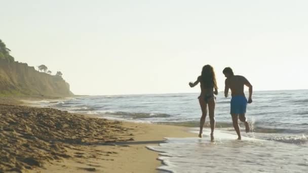 Joyeux jeune couple jouent près du bord de mer dans les vagues de surf sur la plage de sable fin. Mouvement lent — Video