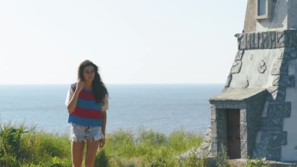 Молодая красивая девушка с распущенными волосами, прогуливающаяся возле здания романтического маяка в солнечный день. Морской пейзаж на заднем плане — стоковое видео