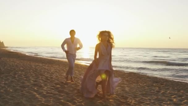 Pareja romántica joven - chica bonita en vestido largo y hombre guapo juguetonamente corre a lo largo de la costa en la playa de arena — Vídeo de stock