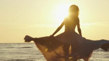 Uzun hafif pembe elbiseli genç çekici kız çevresinde kumlu plaj enlighted üzerinde altın günbatımı ışınları tarafından swirls