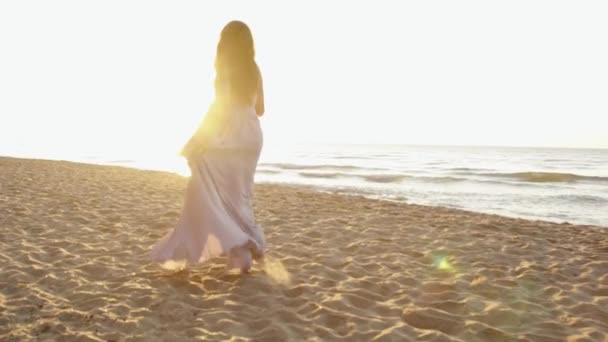 Menina encantadora jovem em vestido de luz longa brinca ao longo da costa na praia de areia e olhando ao redor — Vídeo de Stock