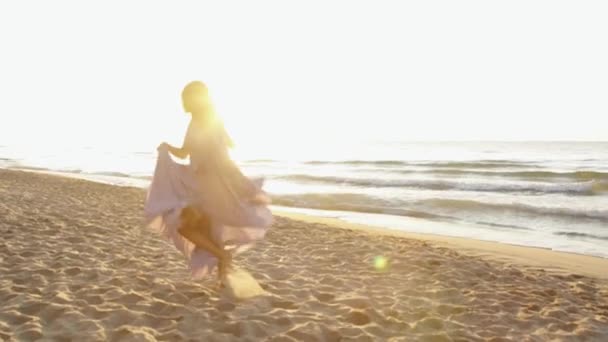 Jeune fille charmante court ludique le long de la côte sur la plage de sable et agitant sa longue robe rose clair — Video