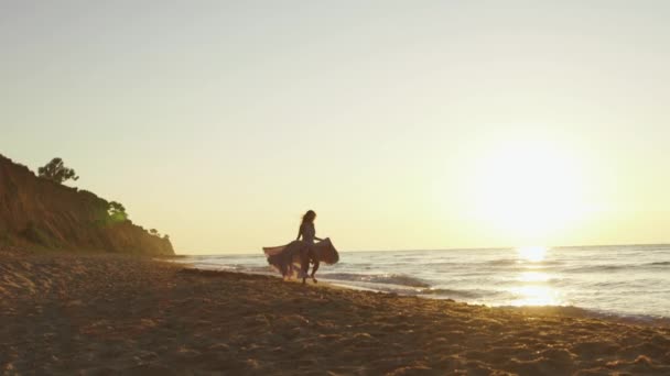 Jeune fille charmante en robe longue et légère court le long de la côte sur une plage de sable éclairée par les rayons dorés du coucher du soleil — Video