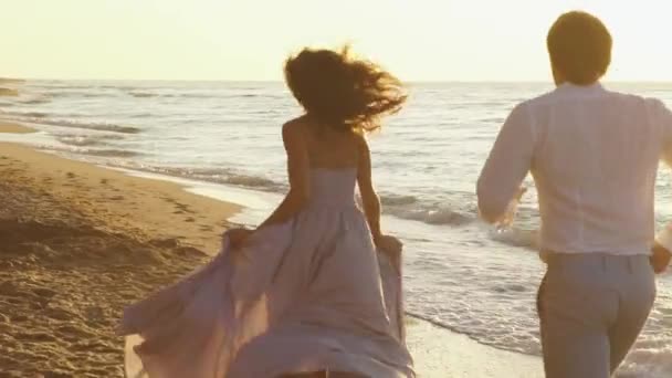 Молодая романтическая пара - красивая девушка в длинном светлом розовом платье и красивый мужчина игриво бегает вдоль побережья на песчаном пляже — стоковое видео