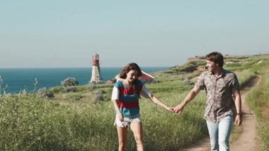 Genç çift holging eller birlikte, konuşmak ve deniz kıyısı cliff fenerinde yakınındaki kum izinde gülerek yürür. Ağır çekim