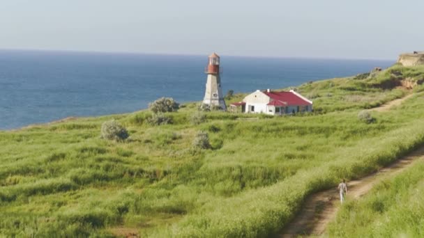 Barfüßiger junger Mann, der an sonnigen Tagen auf einem Sandweg in der Nähe des Leuchtturms spaziert. Meereslandschaft am Horizont — Stockvideo