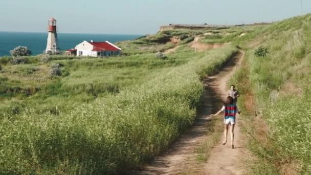 Pareja joven corre uno hacia el otro, se encuentra en el sendero de arena cerca del faro en el acantilado de la orilla del mar — Vídeo de stock