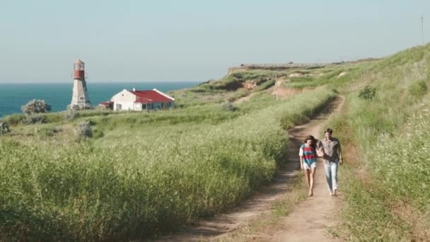 Casal jovem romântico caminha de mãos dadas, conversando e rindo em trilha de areia perto do farol em beira-mar penhasco. Movimento lento — Vídeo de Stock