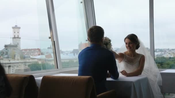 Happy young bride gives to sniff rose bouquet to groom in a cafe with panoramic city view background. — Stock Video