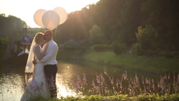 Ömma ögonblick av lycka. Unga vackra bröllopsparet med ballonger omfattar på bakgrund av floden i solnedgången — Stockvideo